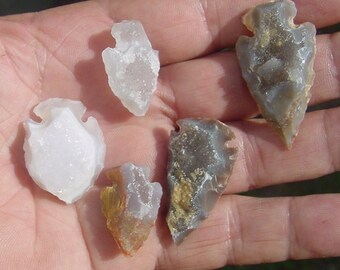 May include: Five arrowheads made of different types of stone. The arrowheads are various shades of gray, white, and brown. Some of the arrowheads have a translucent quality.