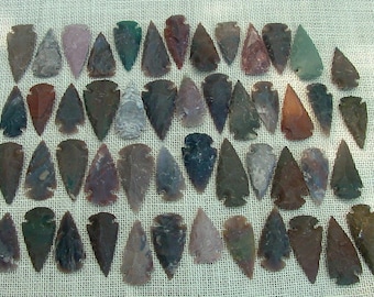 May include: A collection of 48 arrowheads made of various types of stone, including agate, jasper, and flint. The arrowheads are arranged in rows on a beige fabric background. They are all different shapes and sizes, and some have a reddish-brown color.