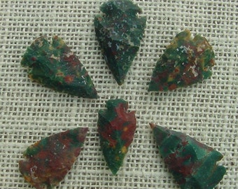 May include: Six arrowheads made of green and red jasper. The arrowheads are arranged in a circle on a beige fabric background.