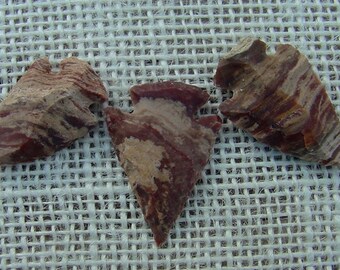May include: Three arrowheads made of red and brown banded stone. The arrowheads are on a white woven fabric background.
