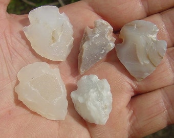 May include: Five pale, translucent arrowhead-shaped stones. The stones vary in color from off-white to light gray, with some showing subtle banding. The edges are rough and irregular, suggesting they are hand-crafted or natural formations. The arrowheads are displayed in an open hand.