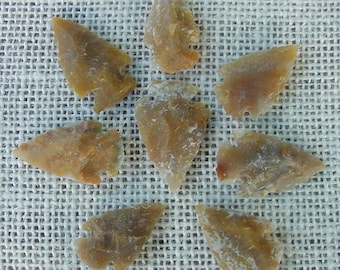 May include: A collection of eight brown arrowheads, some with a white patina, arranged in a star shape on a beige woven fabric background.