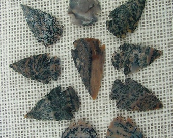 May include: Nine arrowheads made of a mottled, pink, brown, and black stone. The arrowheads are arranged in a circle on a beige fabric background.