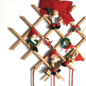 May include: A wooden Christmas tree ornament holder with a red bow and four small felt gnome ornaments hanging from it. The holder has a grid pattern and is decorated with bells and a small wreath.