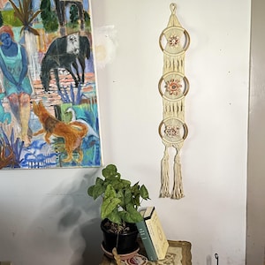 May include: A cream-colored macrame wall hanging with three circular wooden frames, each adorned with a floral design. The macrame features a knotted pattern and tassels at the bottom. The wall hanging is displayed against a white wall.