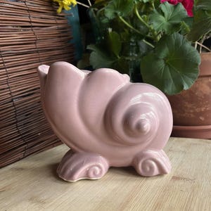 May include: A pink ceramic seashell planter. The planter is shaped like a seashell with a spiral design. It has a smooth, glossy finish.