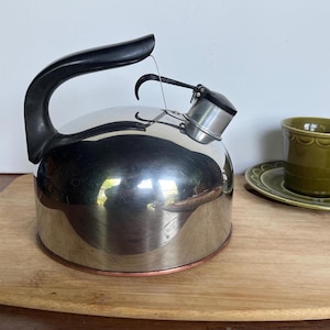May include: A stainless steel tea kettle with a black handle and spout. The kettle sits on a wooden cutting board, with a green teacup and saucer in the background. The kettle has a copper-colored base.