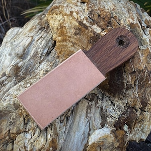 K&ouml;nnte beinhalten: Ein Holz-Lederriemen mit einer Lederscheide. Der Riemen hat einen dunkelbraunen Holzgriff mit einem Loch zum Aufh&auml;ngen. Die Lederscheide ist hellbraun. Der Riemen liegt auf einem gro&szlig;en, strukturierten Felsen.