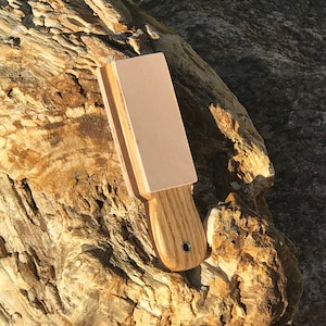 Puede incluir: Una piedra de afilar de cuero marrón claro con un mango de madera. La piedra de afilar está apoyada en un trozo de madera petrificada.