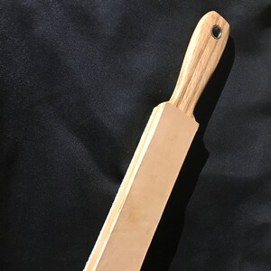 Puede incluir: Una correa de cuero marr&oacute;n claro con un mango de madera. El mango tiene un ojal de metal.