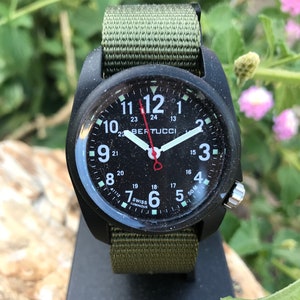 May include: A black and green military style watch with a black face and white numbers. The watch face also has the text "BERTUCCI" and "50M" printed on it. The watch is on a black stand.