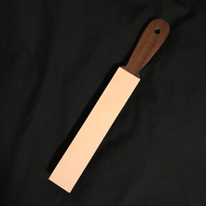 Puede incluir: Una correa de cuero marr&oacute;n claro con un mango de madera marr&oacute;n oscuro. El mango tiene un anillo de metal.