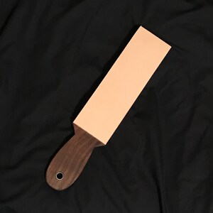 Puede incluir: Una correa de cuero marrón claro con un mango de madera marrón oscuro. La correa se utiliza para afilar navajas de afeitar.