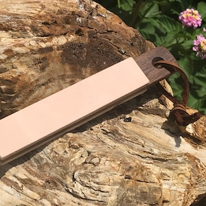 Puede incluir: Una correa de cuero rosa con un mango de madera oscura y una correa de cuero marrón. La correa está apoyada en un trozo de madera petrificada.