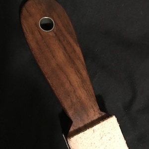 Puede incluir: Una estropa de madera con una superficie de afilado de cuero y un agujero de metal en el mango. La estropa se utiliza para afilar navajas de afeitar.