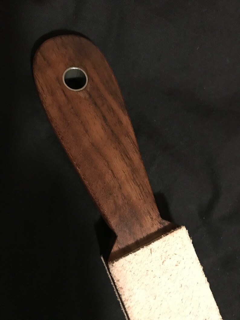 Puede incluir: Una estropa de madera con una superficie de afilado de cuero y un agujero de metal en el mango. La estropa se utiliza para afilar navajas de afeitar.