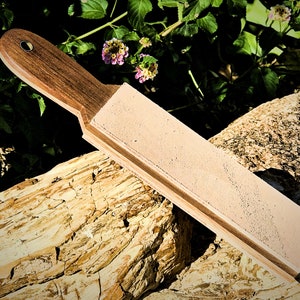 Puede incluir: Una correa de cuero marrón con un mango de madera. La correa está apoyada en un trozo de madera desgastada.