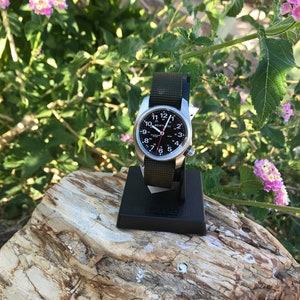 Pu&ograve; includere: Un orologio da polso argentato con quadrante nero e cinturino in nylon verde. L'orologio ha indici bianchi per i numeri e una lancetta dei secondi rossa. L'orologio &egrave; su un supporto nero.