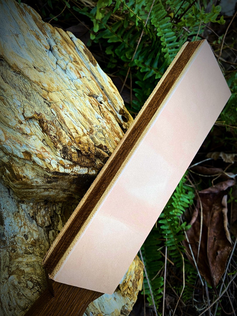 May include: A light pink leather strop with a dark brown wooden handle. The strop is resting on a piece of weathered wood.