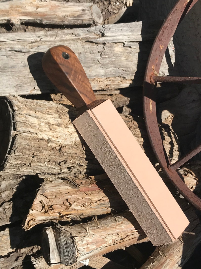 Peut inclure: Une strop en cuir marron avec une poign&eacute;e en bois. La strop a une surface en cuir marron fonc&eacute; avec une bande en cuir marron clair qui descend au centre. La strop repose sur une pile de bois.