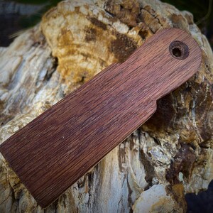 K&ouml;nnte beinhalten: Ein rechteckiger Holzgegenstand mit dunkelbraunem Finish und einem Loch nahe einem Ende. Die Holzmaserung ist sichtbar. Das Objekt, m&ouml;glicherweise ein Werkzeug oder Zubeh&ouml;r, liegt auf einer strukturierten, hellen Oberfl&auml;che.