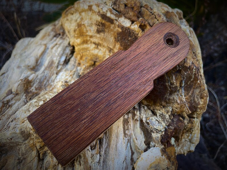 K&ouml;nnte beinhalten: Ein rechteckiger Holzgegenstand mit dunkelbraunem Finish und einem Loch nahe einem Ende. Die Holzmaserung ist sichtbar. Das Objekt, m&ouml;glicherweise ein Werkzeug oder Zubeh&ouml;r, liegt auf einer strukturierten, hellen Oberfl&auml;che.