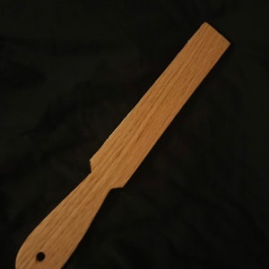 May include: A wooden paddle with a rounded handle and a hole in the handle. The paddle is light brown in colour and has a smooth finish.