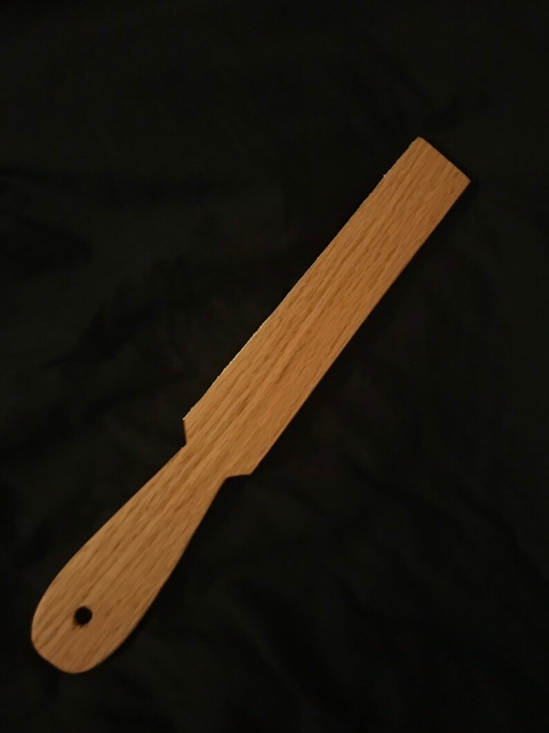 May include: A wooden paddle with a rounded handle and a hole in the handle. The paddle is light brown in colour and has a smooth finish.