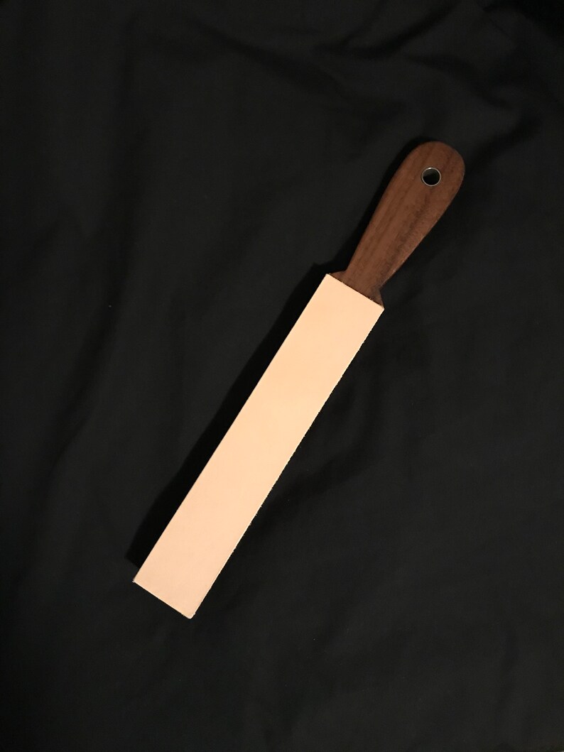 Puede incluir: Una correa de cuero marr&oacute;n claro con un mango de madera marr&oacute;n oscuro. El mango tiene un anillo de metal.