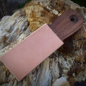 K&ouml;nnte beinhalten: Ein Holzriemen mit einer Lederscheide. Der Riemen besteht aus dunkelbraunem Holz und hat ein Loch am Ende. Die Scheide ist hellrosa und bedeckt die Klinge des Riemens. Der Hintergrund ist ein strukturierter Felsen.