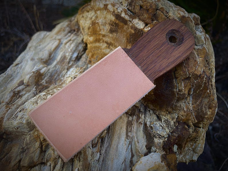 K&ouml;nnte beinhalten: Ein Holzriemen mit einer Lederscheide. Der Riemen besteht aus dunkelbraunem Holz und hat ein Loch am Ende. Die Scheide ist hellrosa und bedeckt die Klinge des Riemens. Der Hintergrund ist ein strukturierter Felsen.