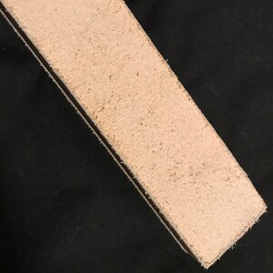 Puede incluir: Primer plano de una correa de cuero beige claro con textura rugosa. La correa est&aacute; sobre una superficie negra.