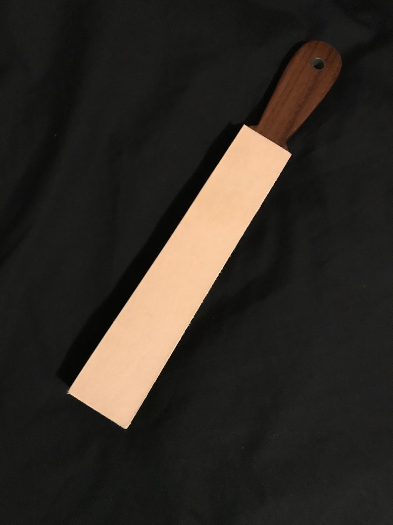 Puede incluir: Una correa de cuero marr&oacute;n claro con un mango de madera marr&oacute;n oscuro. La correa se utiliza para afilar navajas de afeitar.
