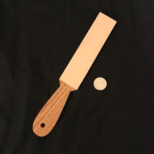 Puede incluir: Un mango de madera marrón claro con un agujero en el centro unido a una tira de cuero rosa claro. La tira mide aproximadamente 25 cm de largo y 5 cm de ancho.