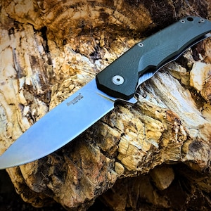 May include: A green and silver folding knife with a black handle and a partially open blade. The knife is resting on a piece of weathered wood.