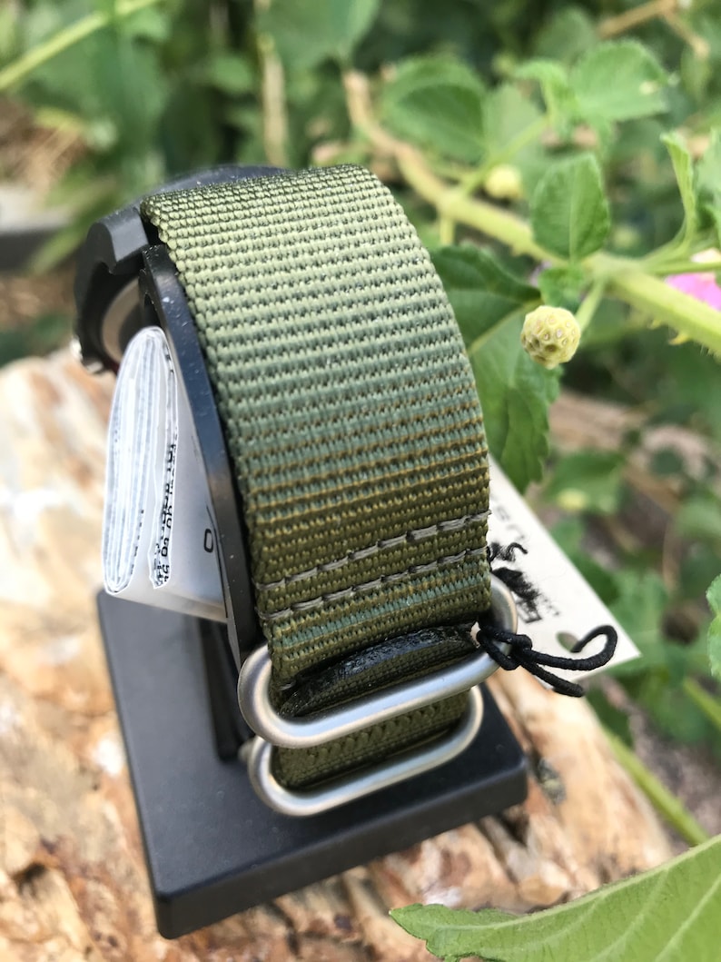 May include: A green and brown nylon watch strap with silver metal hardware. The strap is attached to a watch face that is not visible in the image.