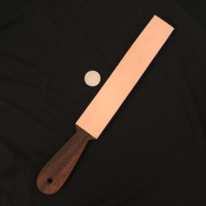 Puede incluir: Una correa de cuero rosa claro con un mango de madera marrón oscuro. La correa mide aproximadamente 30 cm de largo y 5 cm de ancho. Se utiliza para afilar navajas de afeitar.