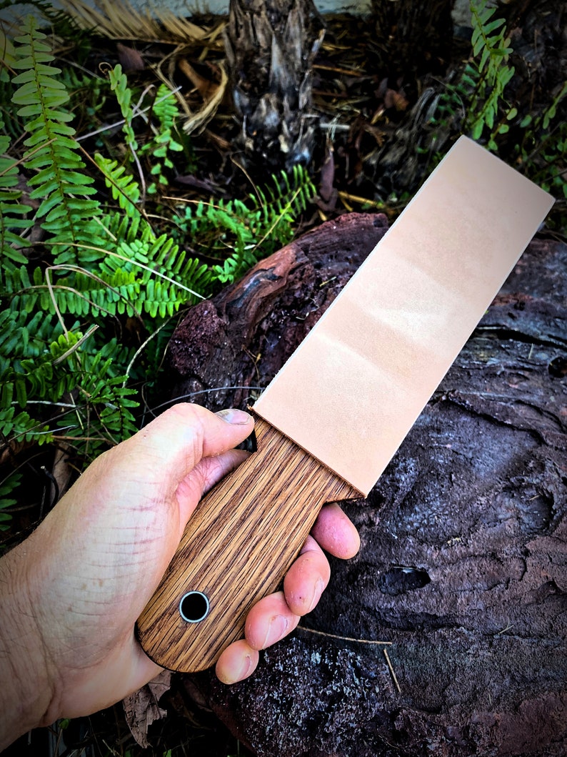 May include: A wooden handled leather strop for sharpening knives. The strop has a light brown leather surface and a dark brown wooden handle with a metal ring.