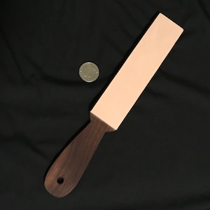 May include: A light pink leather strop with a dark brown wooden handle. The strop is used for sharpening straight razors.