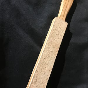 Puede incluir: Una correa de cuero con un mango de madera. La correa es beige y el mango es marr&oacute;n claro con un patr&oacute;n de grano. El mango tiene un anillo de metal en la parte superior.
