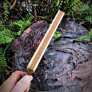 May include: A wooden sharpening steel with a light brown finish. The steel is held in a hand against a dark brown, textured background.