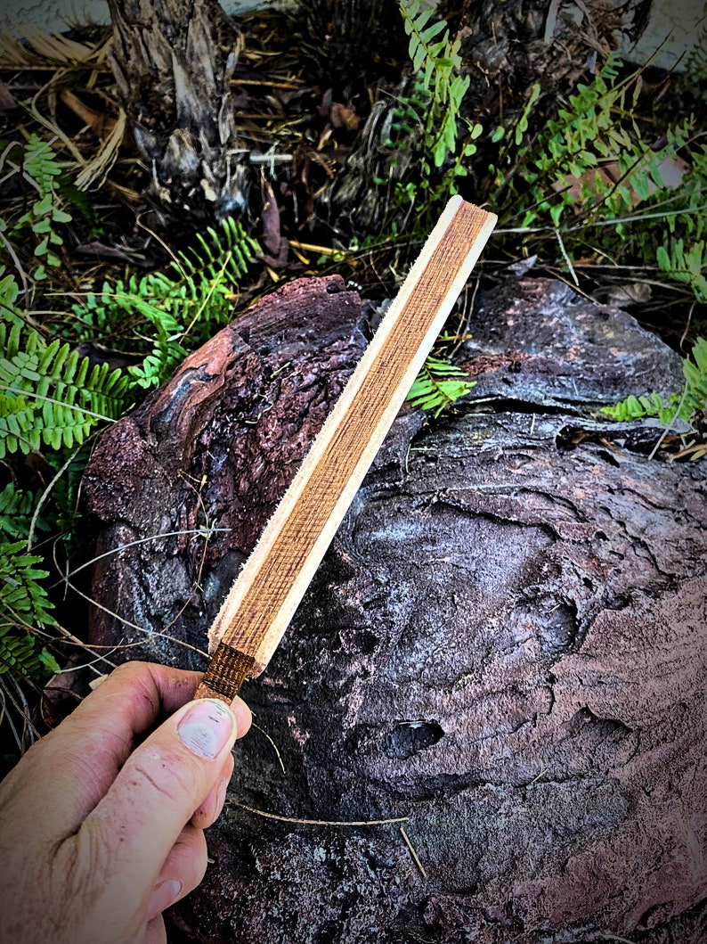 May include: A wooden sharpening steel with a light brown finish. The steel is held in a hand against a dark brown, textured background.