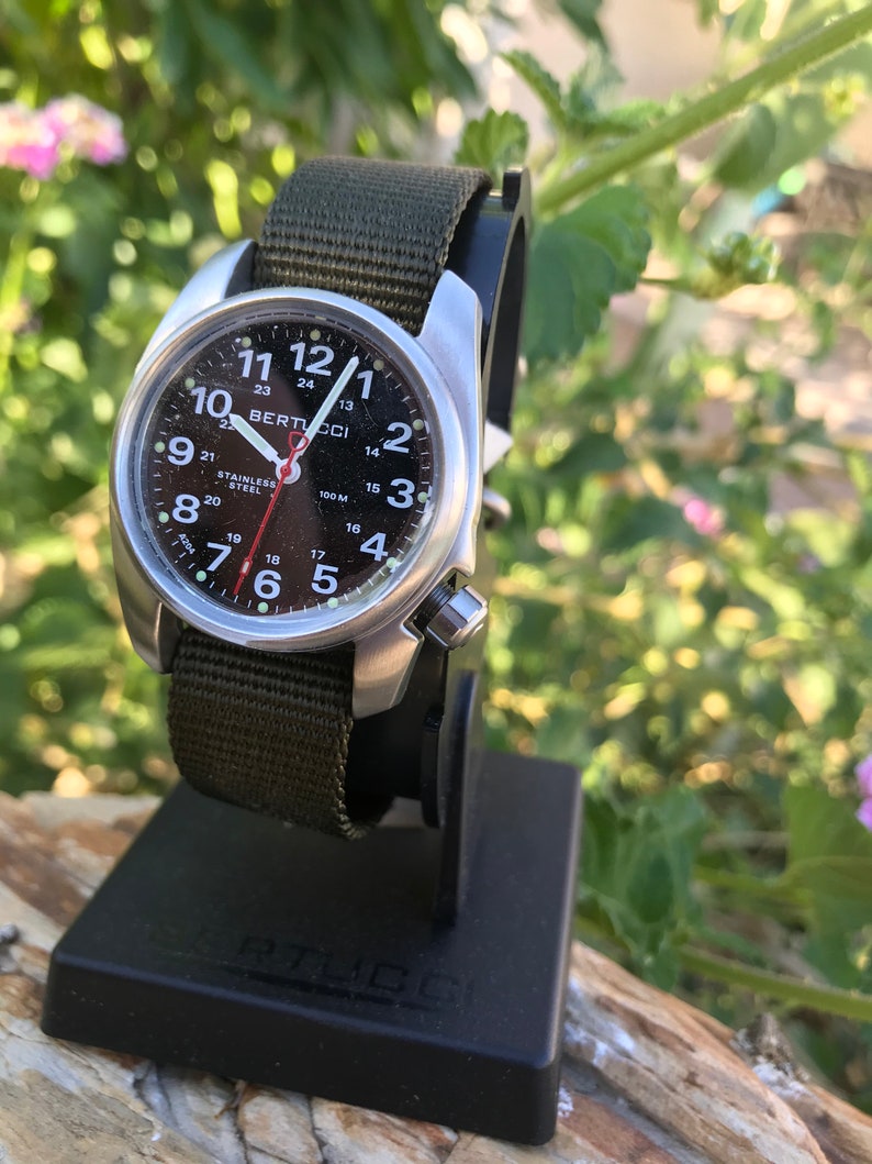 Pu&ograve; includere: Un orologio Bertucci nero e argento con un cinturino in nylon verde. Il quadrante dell'orologio ha numeri e lancette bianchi, e una lancetta dei secondi rossa. L'orologio &egrave; su un supporto per orologio nero.