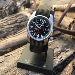 Pu&ograve; includere: Un orologio da polso argentato con quadrante nero e cinturino in nylon verde. Il quadrante dell'orologio ha numeri e segni bianchi, e il nome del marchio "BERTUCCI" &egrave; stampato in nero. L'orologio &egrave; su un supporto nero con il nome del marchio "BERTUCCI" stampato in bianco.