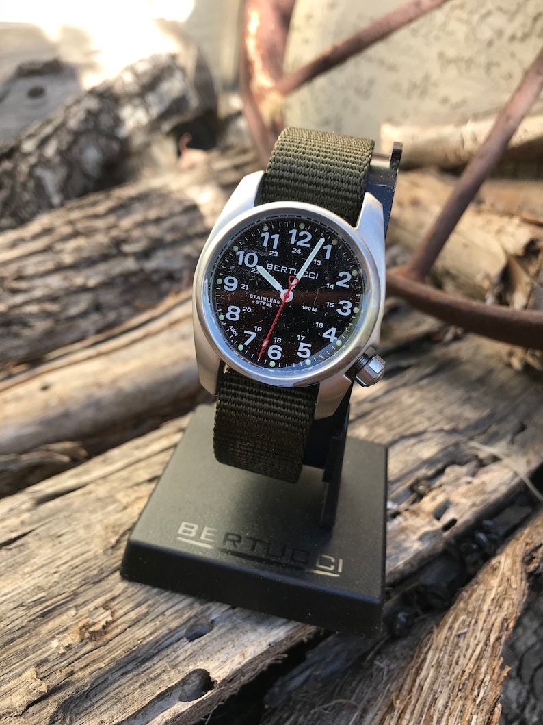Pu&ograve; includere: Un orologio da polso argentato con quadrante nero e cinturino in nylon verde. Il quadrante dell'orologio ha numeri e segni bianchi, e il nome del marchio "BERTUCCI" &egrave; stampato in nero. L'orologio &egrave; su un supporto nero con il nome del marchio "BERTUCCI" stampato in bianco.