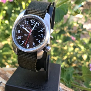 Pu&ograve; includere: Un orologio Bertucci nero e argento con un cinturino in nylon verde. Il quadrante dell'orologio ha numeri bianchi e una lancetta dei secondi rossa. L'orologio &egrave; su un supporto per orologio nero.