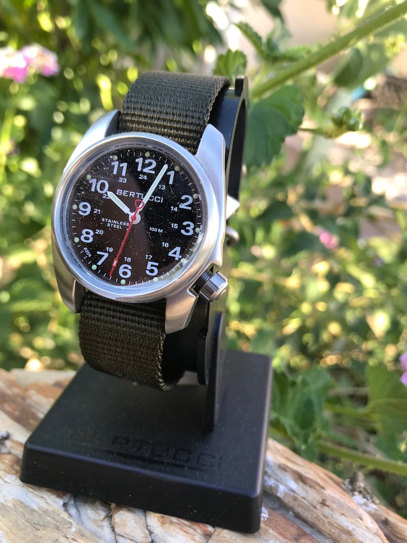 Pu&ograve; includere: Un orologio Bertucci nero e argento con un cinturino in nylon verde. Il quadrante dell'orologio ha numeri bianchi e una lancetta dei secondi rossa. L'orologio &egrave; su un supporto per orologio nero.