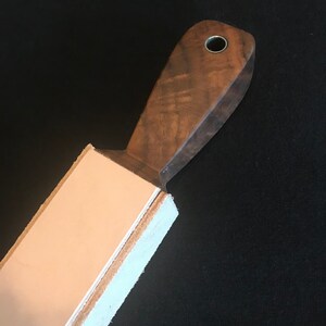 Peut inclure: Une strop en cuir avec une poign&eacute;e en bois. La poign&eacute;e est en bois fonc&eacute; avec un motif de grain naturel. La strop est blanche et a une surface en cuir brun clair.