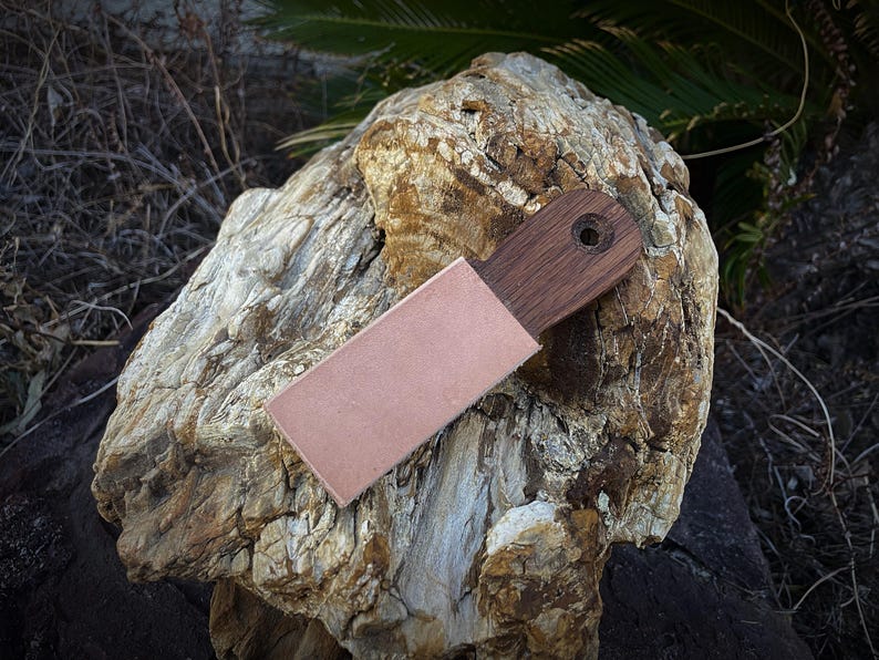 K&ouml;nnte beinhalten: Ein Holz-Lederriemen mit einer Lederscheide. Der Riemen hat einen dunkelbraunen Holzgriff mit einem Loch zum Aufh&auml;ngen. Die Lederscheide ist hellbraun. Der Riemen liegt auf einem gro&szlig;en, strukturierten Felsen.