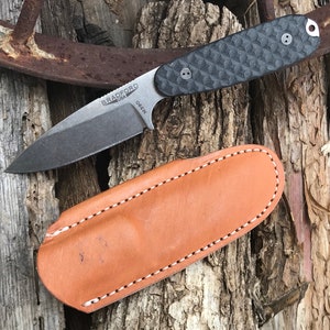 May include: A Bradford USA folding knife with a dark blade and textured black handle, resting on a wooden surface. The knife is accompanied by a brown leather sheath with white stitching.